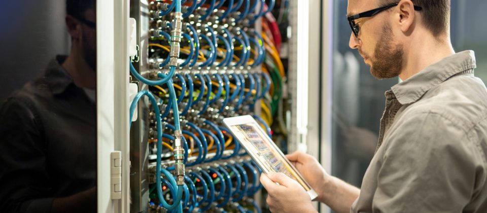 Serious pensive young data center technician in glasses standing against cabinet of server and using digital tablet while checking connections