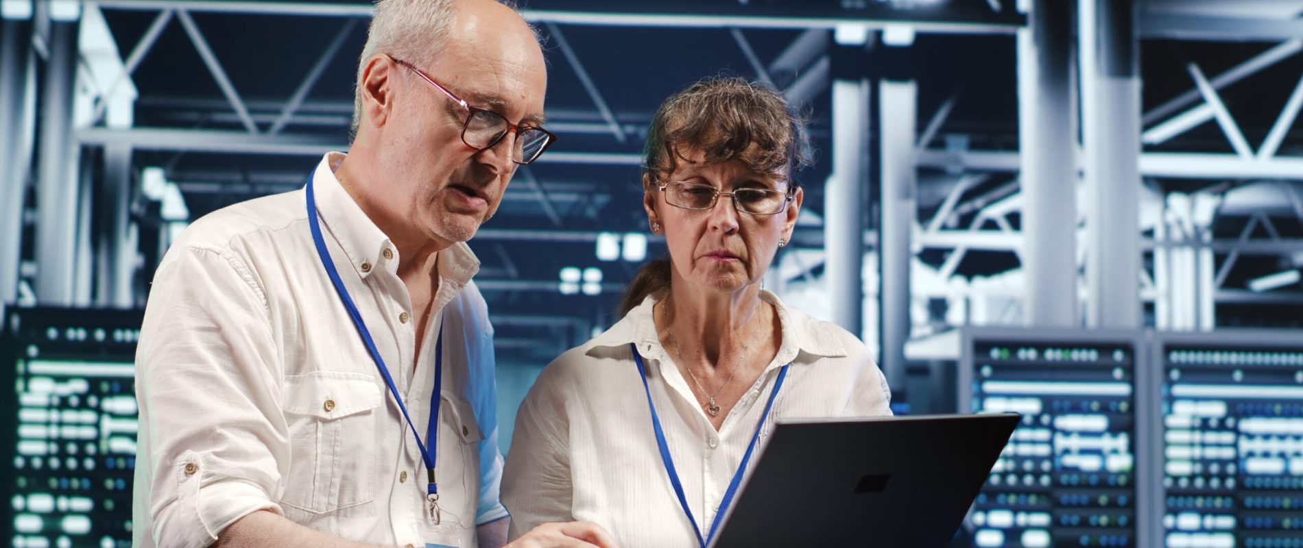 Octogenarian IT team looking around high tech data center, using laptop to crosscheck disaster recovery plan and assess server devices in need of replacement, preventing bugs